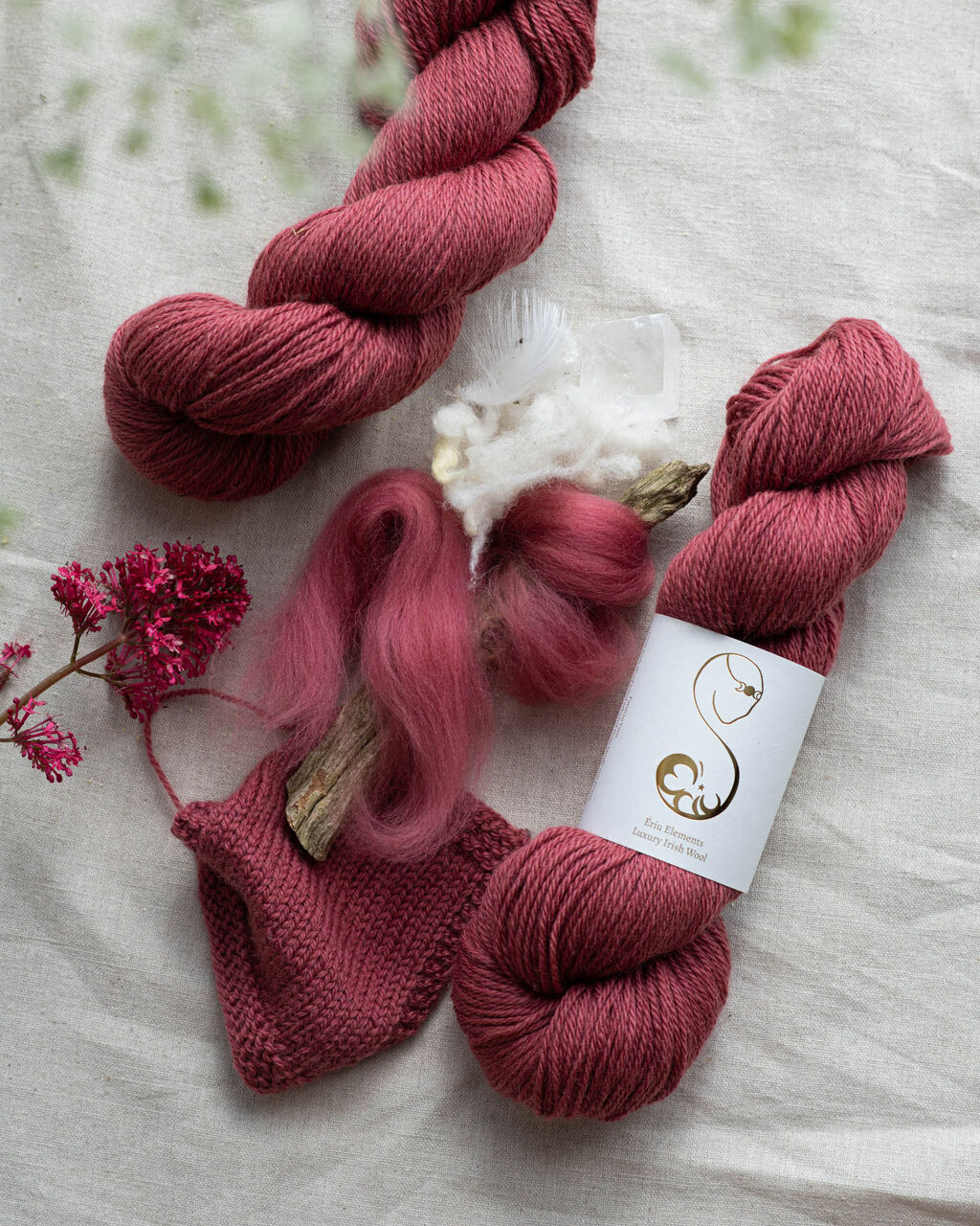 Dark pink yarn skeins with a knitted square and roving decorated with flowers on a light background