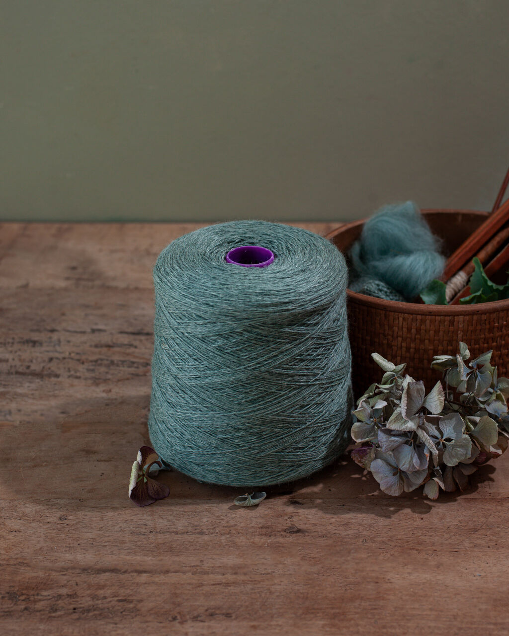 a cone of teal yarn wool on a wooden surface with a plant and basket with teal roving in the background