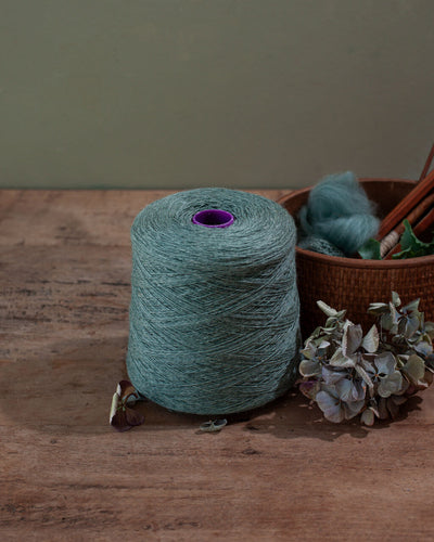 a cone of teal yarn wool on a wooden surface with a plant and basket with teal roving in the background