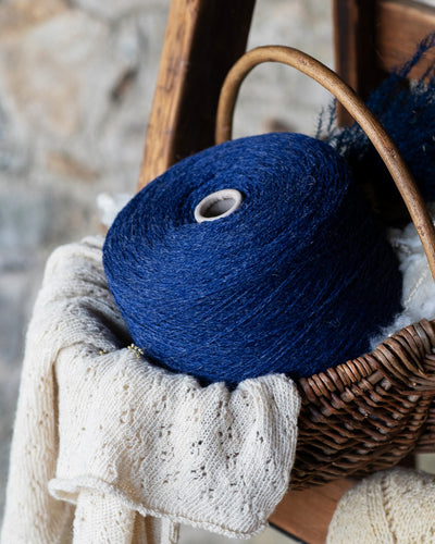 Dark blue colored wool spun on a cone in a wicker basket with raw wool and and natural color knitted shawl