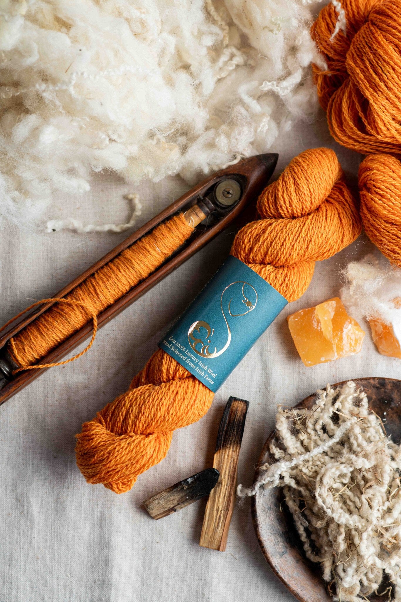 Bright orange wool yarn in a skein, with a boat shuttle, amber decoration, placed on a linen surface.