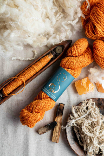 Bright orange wool yarn in a skein, with a boat shuttle, amber decoration, placed on a linen surface.