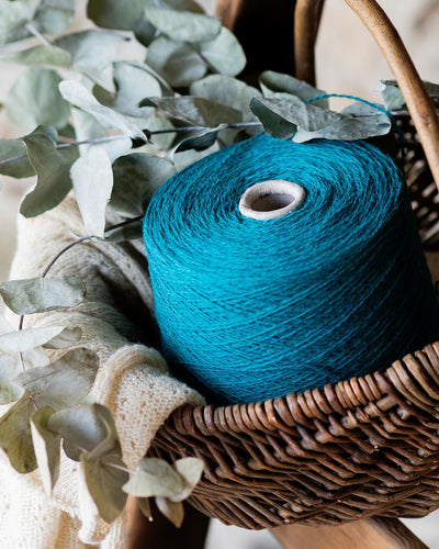 Acone of turquoise wool yarn placed in a brown wicker basket with dull-green foliage and a beige knit fabric.