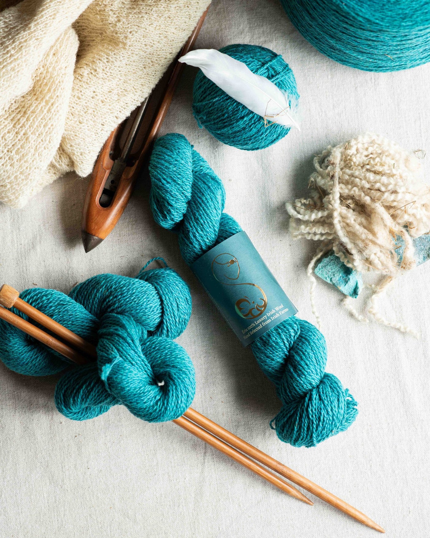 A skein of turquoise wool yarn alongside knitting tools and a partially knitted fabric piece on a white background.