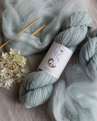 Light blue-gray skein of yarn, knitting needles and floral elements on a wooden surface.
