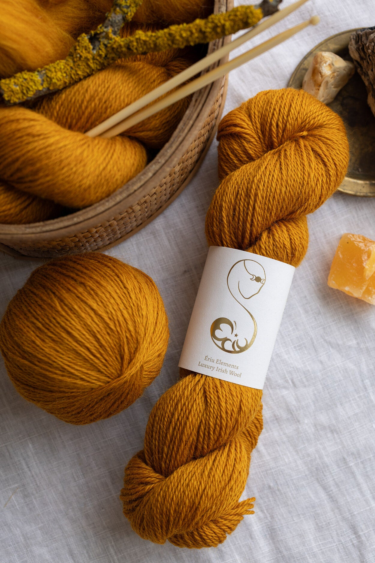 Ochre yarn skein with a label on a light gray surface, next to a basket of yarn and knitting needles.