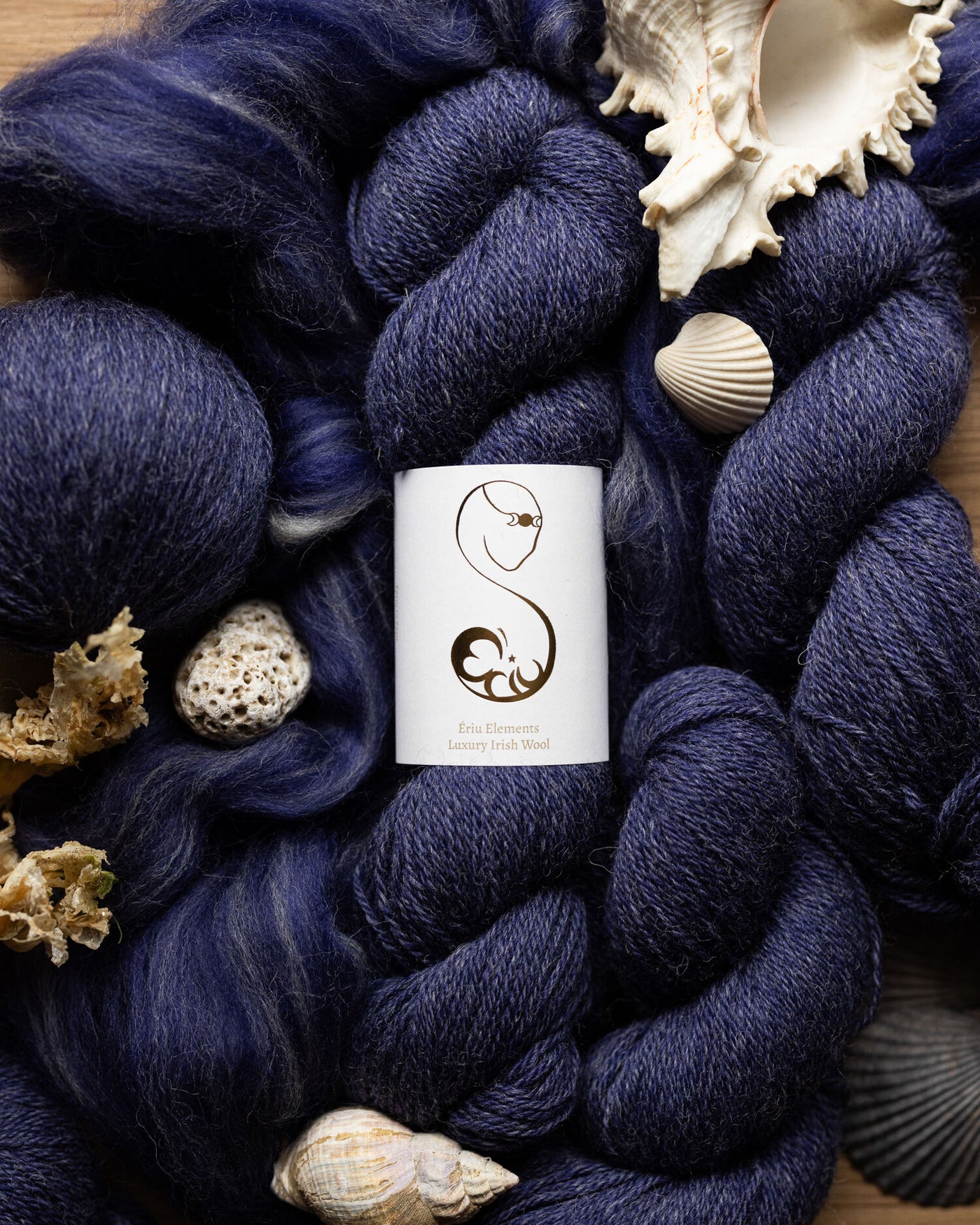 Navy luxury wool skeins with a cardboard product label surrounded by seashells on a wooden surface