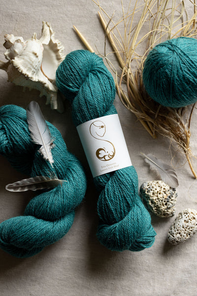 Teal yarn skeins with Ériu label on a textured surface with feathers and shells.
