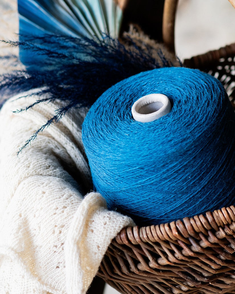A deep turquoise blue cone of yarn in a wicker basket with an uncolored shawl and navy blue feathers