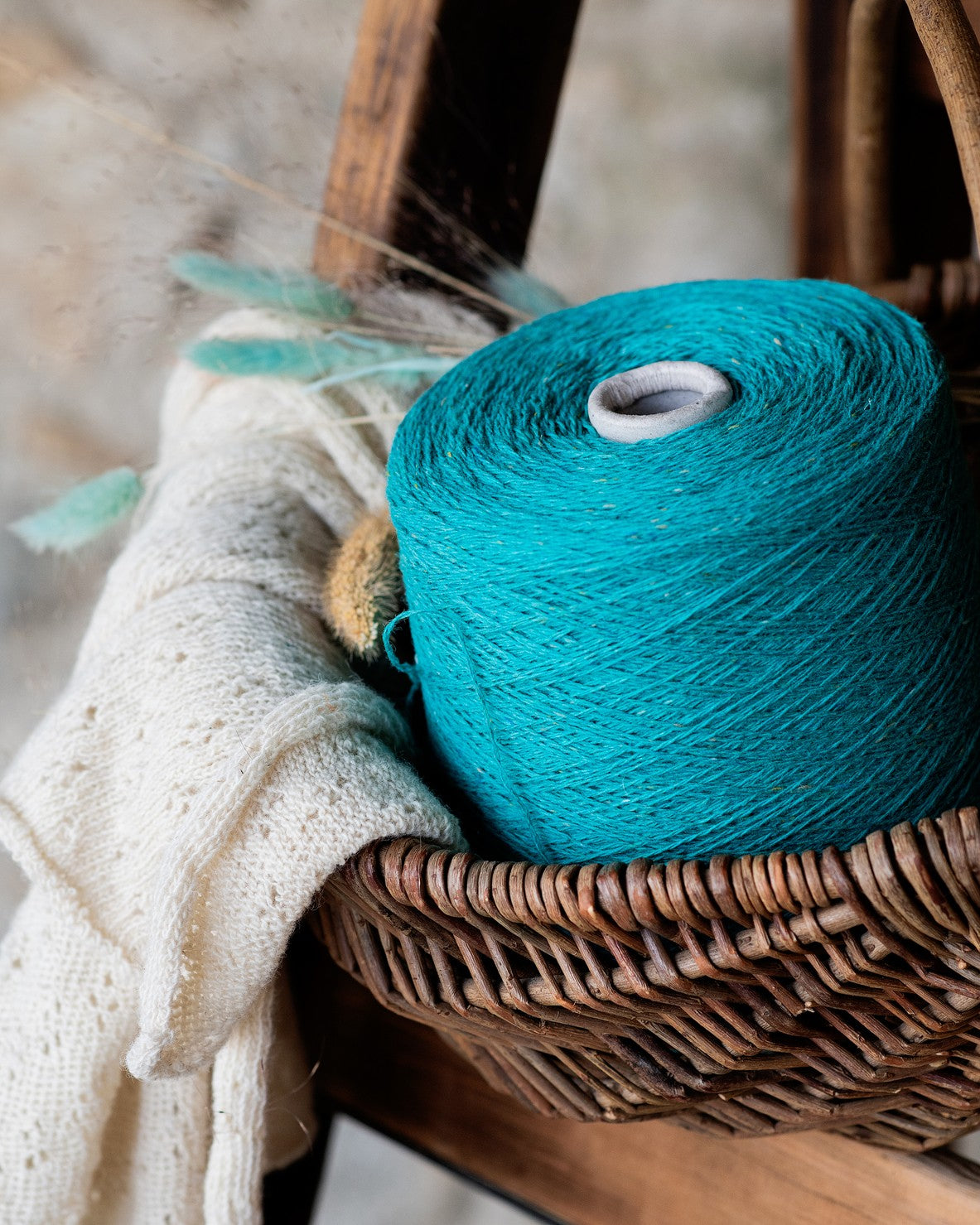A mermaid green wool yarn cone placed in a wicker basket with a knitted piece and a natural-colored scarf