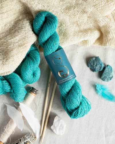 Mermaid blue wool yarn skein on a
light background with branded label,
alongside decorative feathers and a shawl