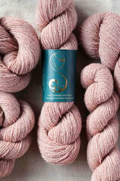 Skeins of light pink wool yarn with a label showing the brand and 100% wool fiber content.