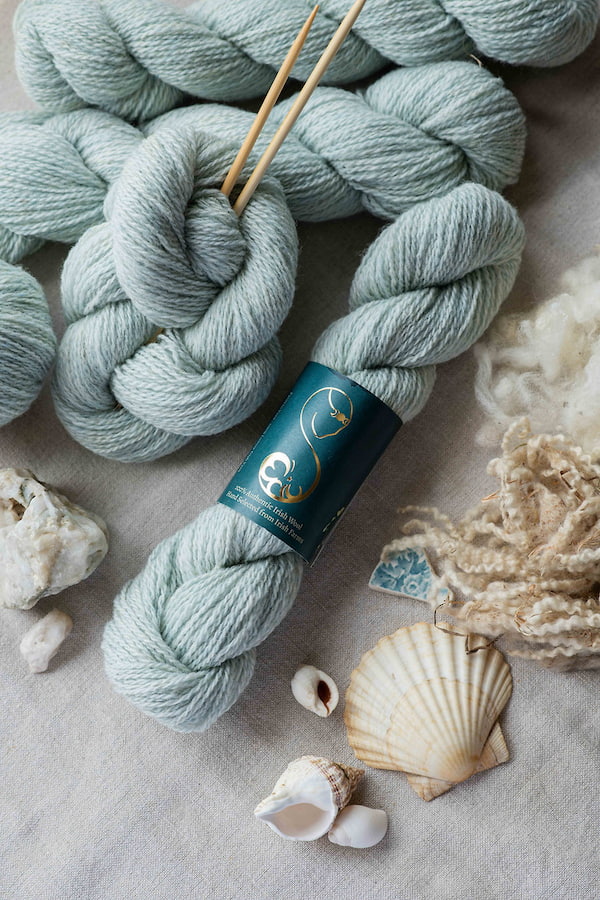 A delicate light green skein of wool yarn, displayed with seashells and raw wool fibers.