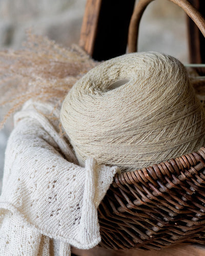 A cone of wool yarn in a sheep's back color, placed in a woven basket, with a knitted scarf and dried wheat decoration