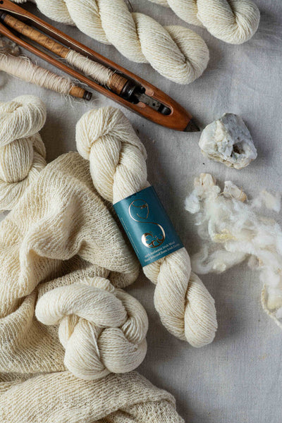 Skeins of off-white wool yarn, one with a turquoise label, decorated with knitting tools and a white stone
